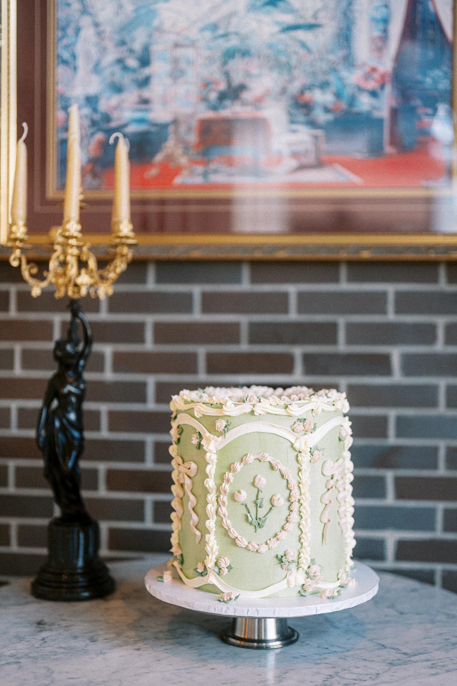 A beautifully decorated green cake with intricate icing designs sits on a cake stand in front of a brick wall, accompanied by an ornate candelabra and artwork. Perfect for elegant dessert or event planning concepts.