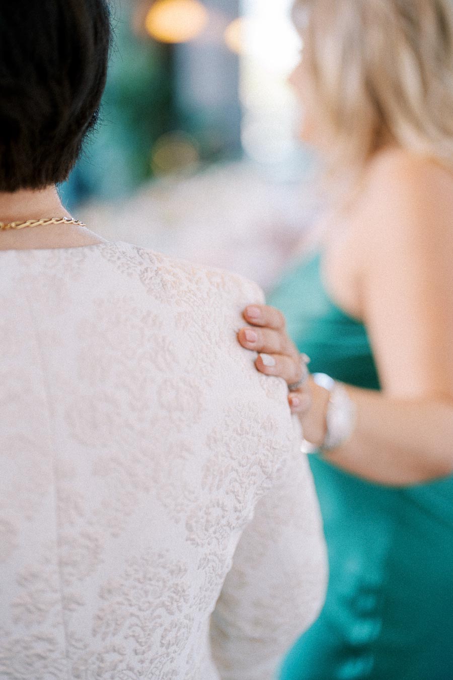 Two people at an event, with one placing a hand on the other's shoulder. The person in the foreground wears a white, textured outfit and a gold necklace, while the person in the background wears a teal dress. The setting suggests a formal or celebratory occasion.