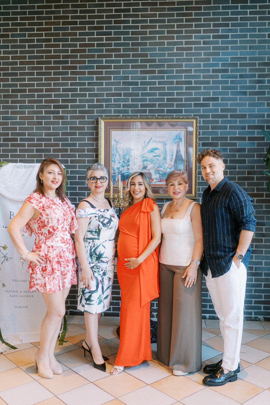 A group of five people smiling and posing at a baby shower, with one individual wearing a bright orange dress that indicates pregnancy. They stand in front of a brick wall with floral decorations and a framed painting in the background.