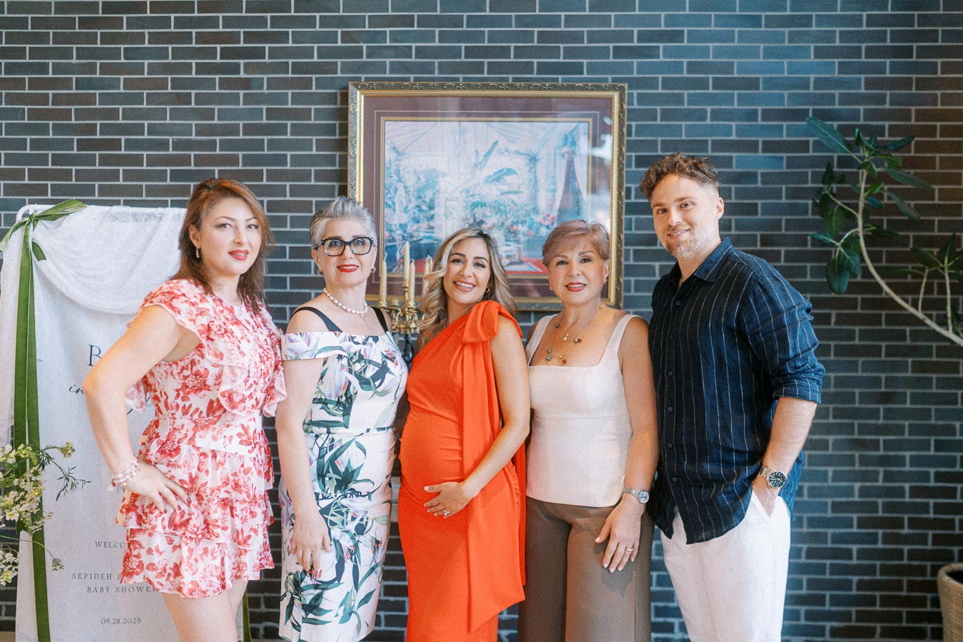 A group of five people smiling at a baby shower, with one woman in a bright orange dress holding her belly, standing against a stylish brick wall backdrop.