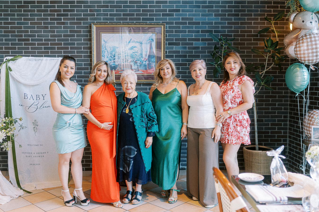 Group of women celebrating a baby shower with Baby in Bloom banner and stylish outfits in a decorated venue.