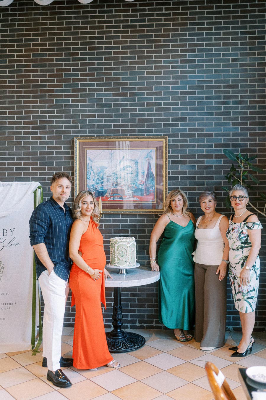 A group of five elegantly dressed people standing around a decorative cake on a small table against a brick wall backdrop, under a framed painting. Celebratory event setting with a mix of colorful outfits.