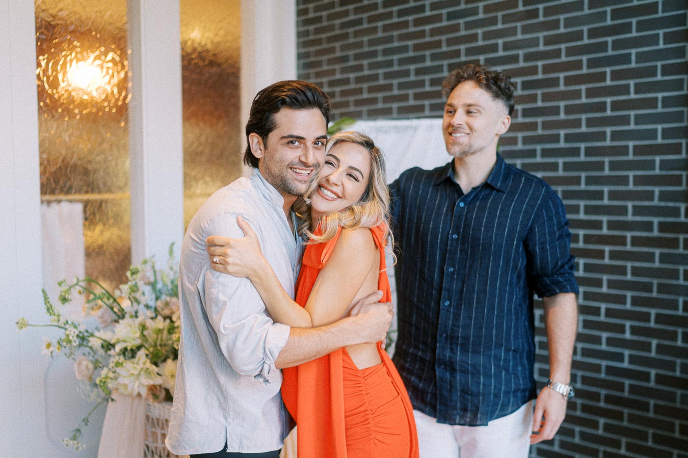 Three people smiling and embracing indoors, with a brick wall and floral arrangement in the background, exuding joy and camaraderie.