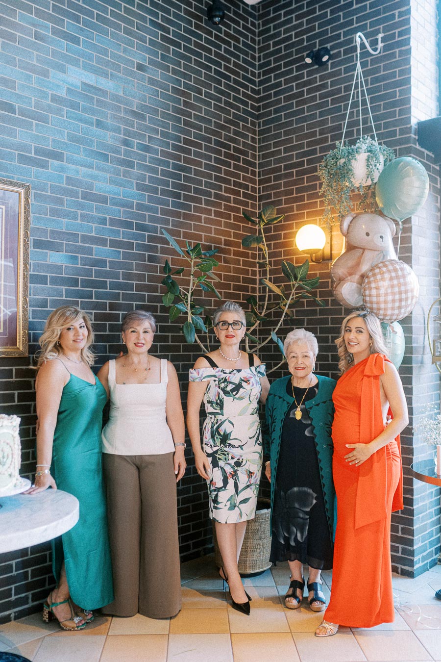 A group of five women in vibrant dresses and outfits stand together in a stylish setting with brick walls and decorative balloons.