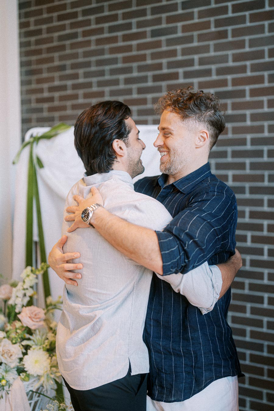 Two men embracing warmly in front of a brick wall, surrounded by flowers, conveying friendship and happiness