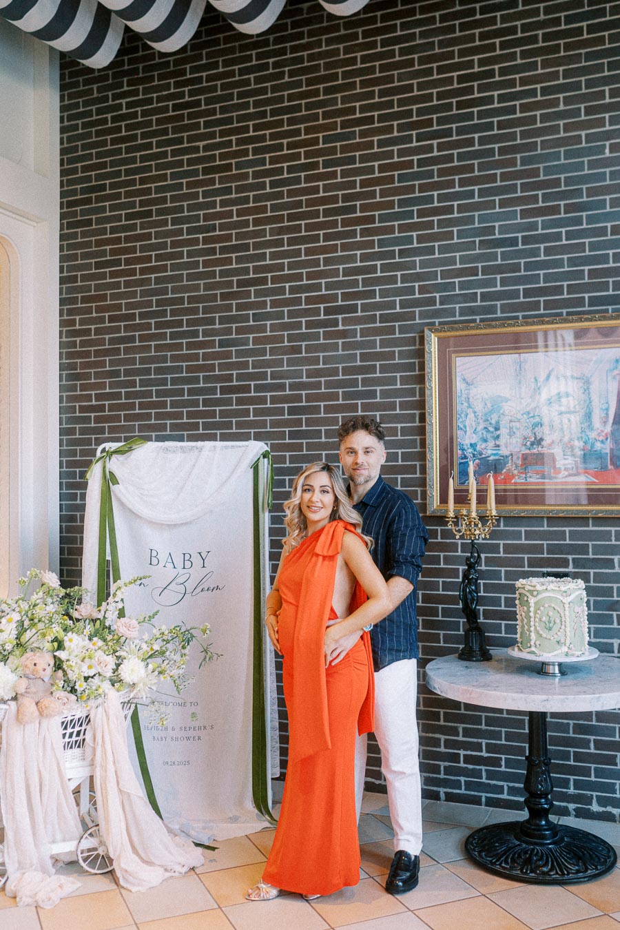 A couple posing in front of a Baby Bloom themed baby shower setup, with elegant floral arrangements and a decorative cake. The woman in an orange dress and the man in a dark shirt are smiling, celebrating their upcoming arrival.