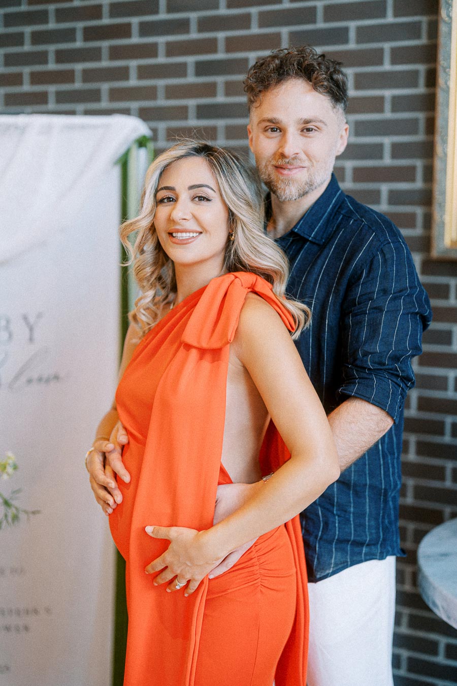 A smiling couple posing together, with the woman in a vibrant orange dress and the man in a blue-striped shirt, standing against a brick wall backdrop.