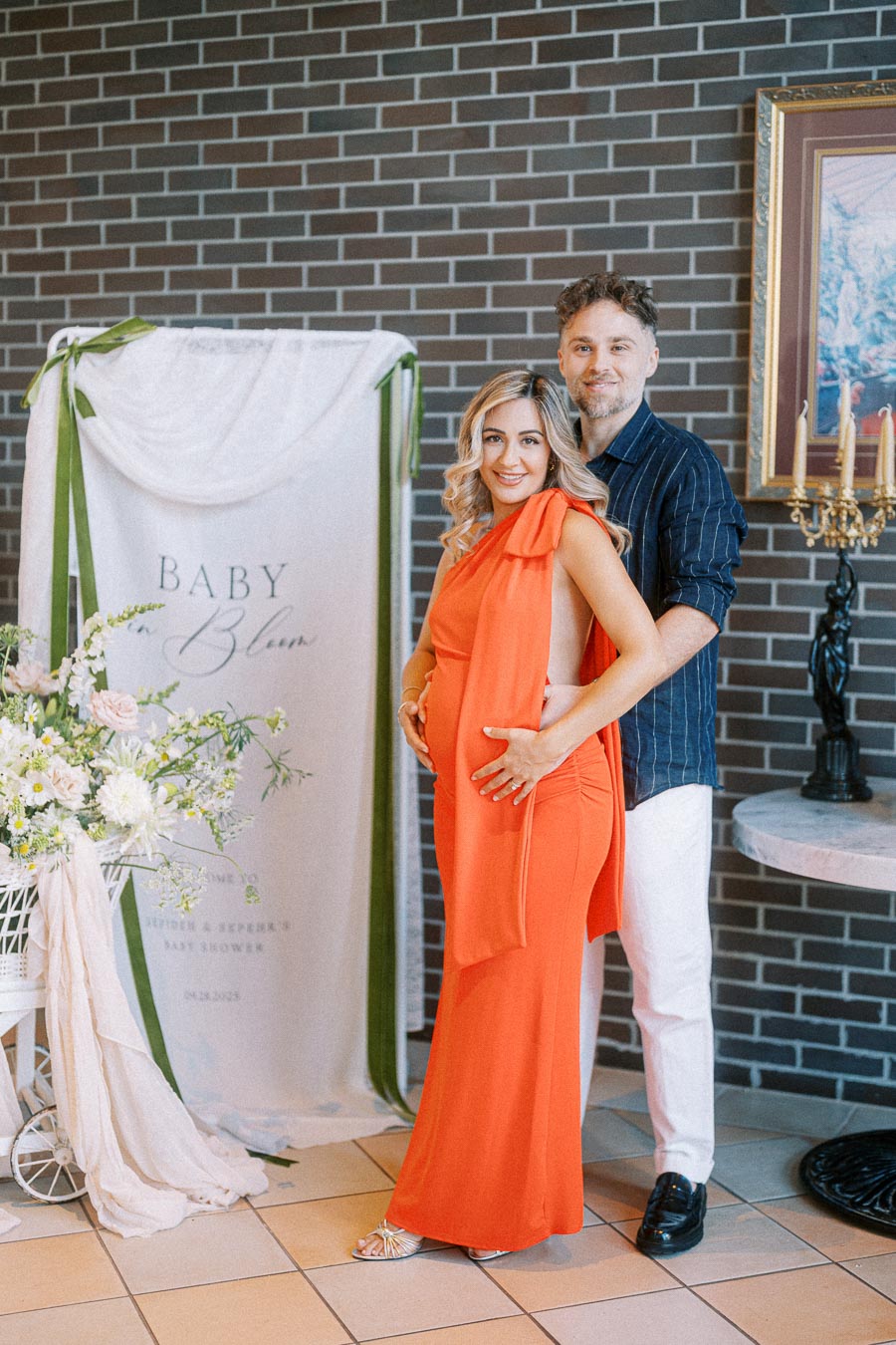 Couple posing at an indoor baby shower, with the woman in an orange dress and the man in a dark shirt. They stand in front of a Baby in Bloom sign surrounded by elegant floral arrangements, highlighting a joyous celebration.