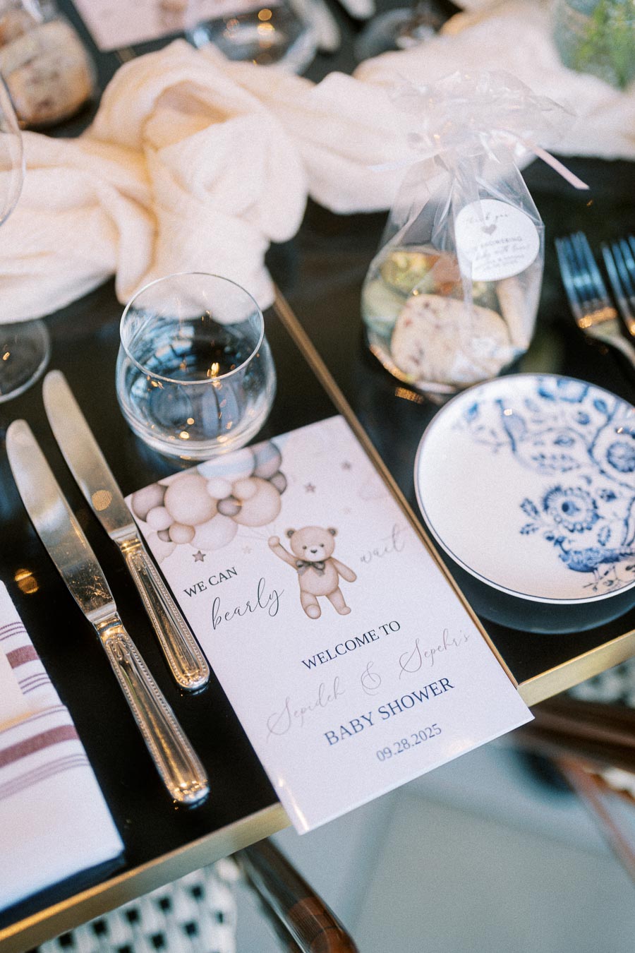 Elegant table setting for a baby shower with a menu featuring a teddy bear and balloon design, flanked by polished cutlery, a glass, a blue floral plate, and a wrapped party favor bag.
