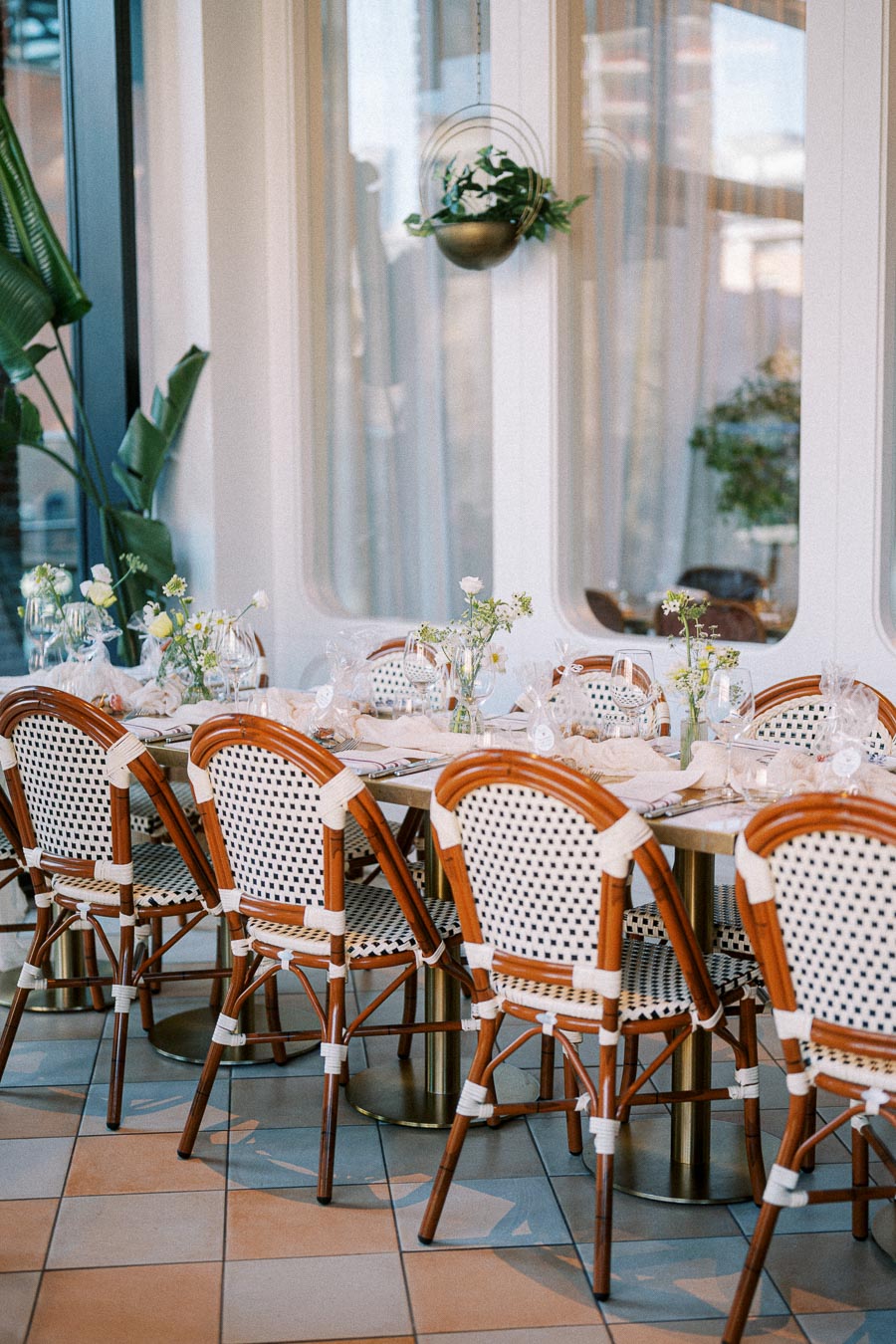 Upscale dining area with elegant table setting for eight, featuring white woven chairs and delicate floral centerpieces, in a bright, modern restaurant with large windows and hanging plants.
