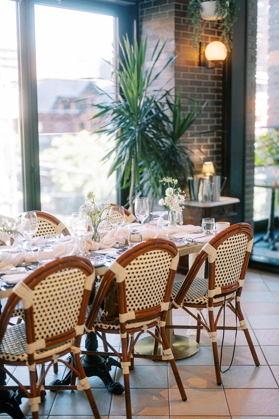 Elegant dining setup with wicker chairs and a decorated table featuring glassware and vases of flowers in a sunlit indoor space near large windows and greenery.