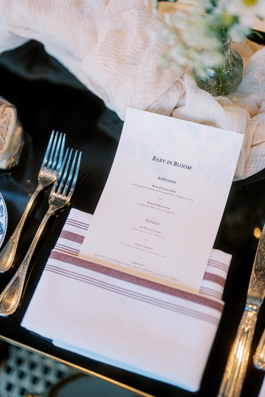 Elegant table setting featuring a Baby in Bloom menu on a striped napkin, accompanied by silver cutlery and a blurred floral arrangement in the background.