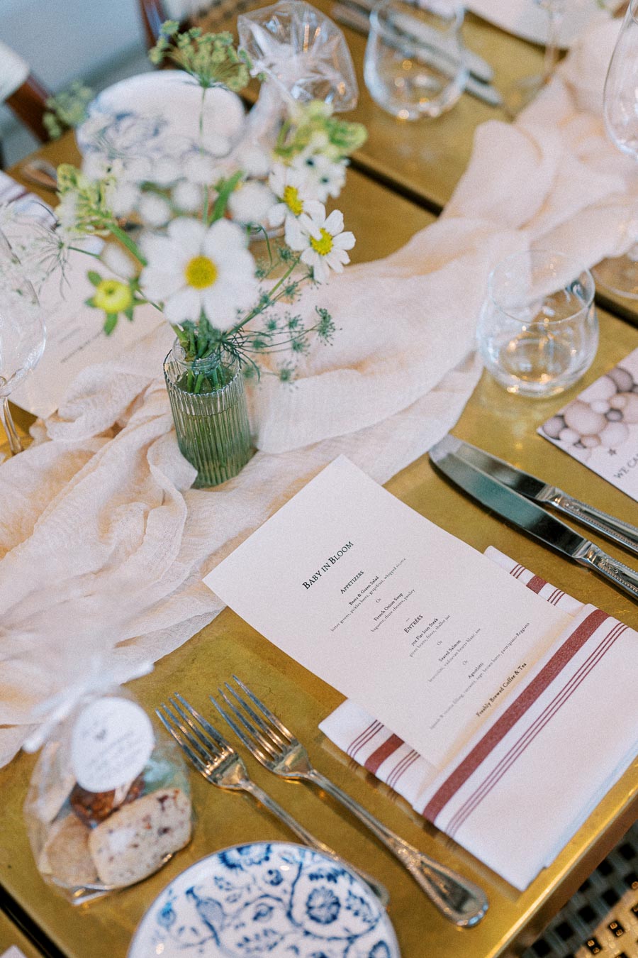 Elegant table setting with a menu card titled Baby in Bloom, featuring delicate white flowers in a glass vase, a white tablecloth, and neatly arranged cutlery on a golden surface.