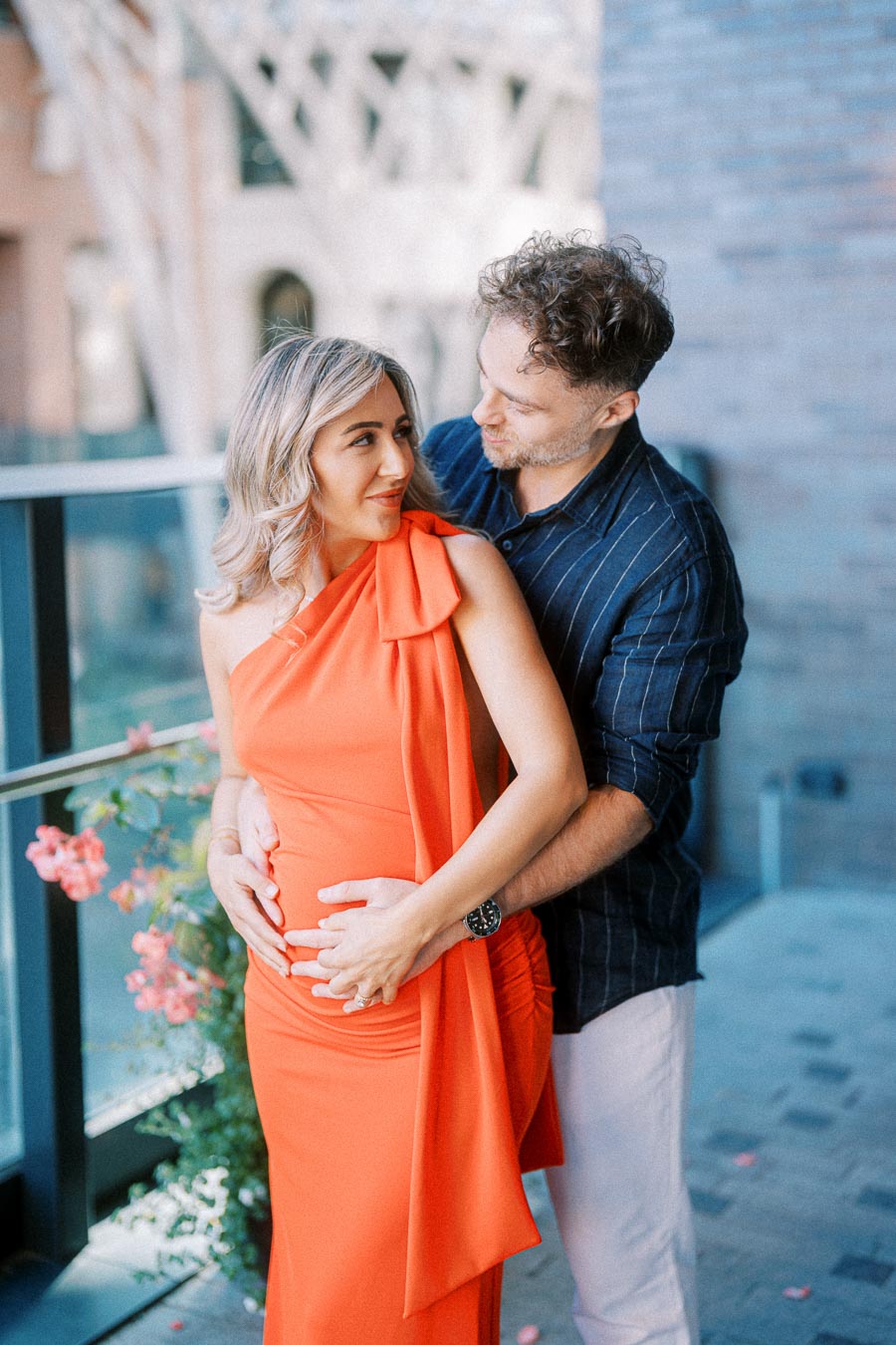 A couple embracing on a balcony; the woman, in a vibrant orange dress, is pregnant, and the man, wearing a dark shirt, lovingly holds her belly outdoors with flowers and a blurred urban background.
