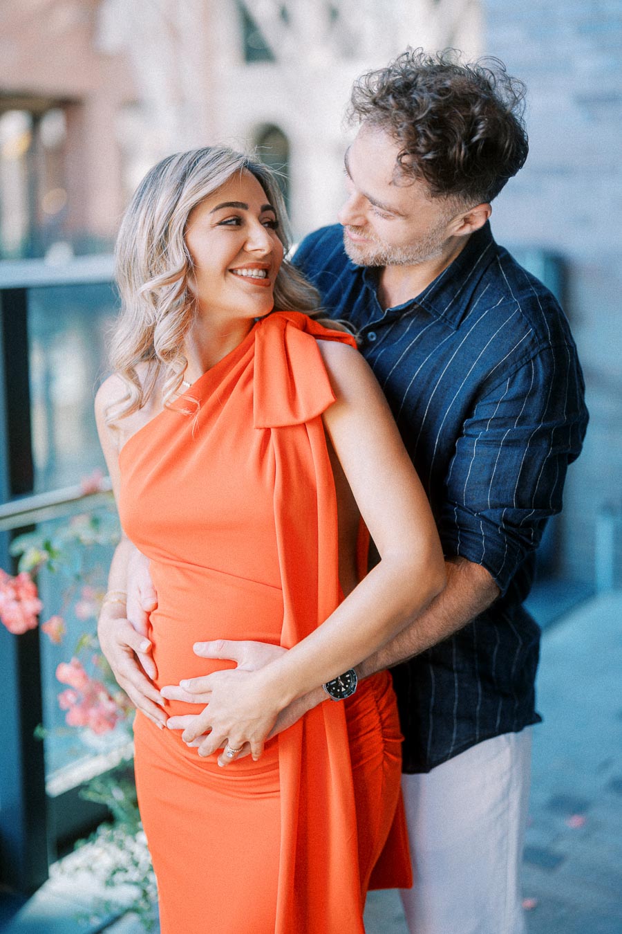 Couple embracing during maternity photoshoot, woman wearing an orange dress and man in a striped shirt, outdoors.