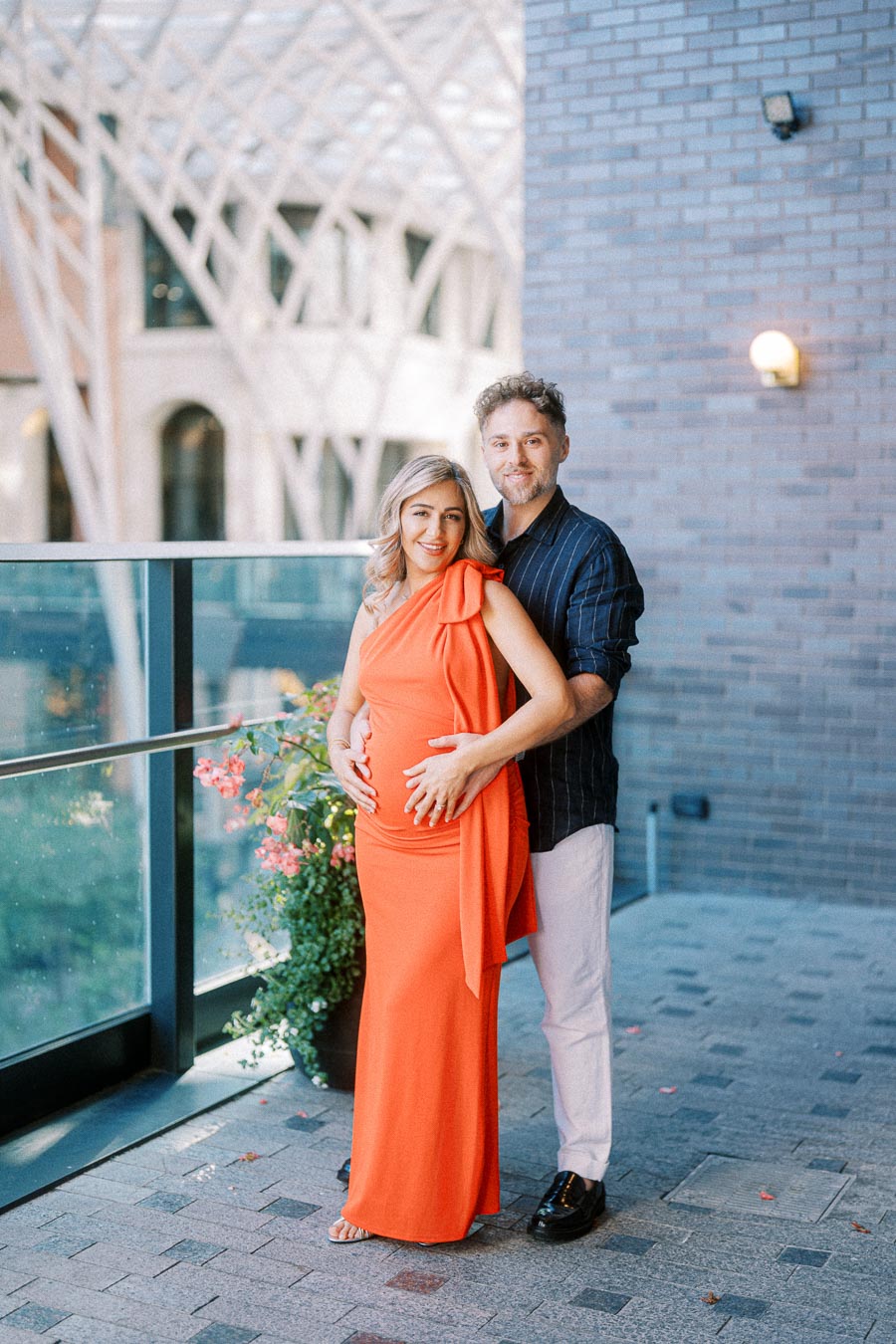 A couple embracing joyfully on a terrace, with the woman wearing an elegant orange dress and holding her pregnant belly, framed by a modern architectural backdrop and blooming flowers.
