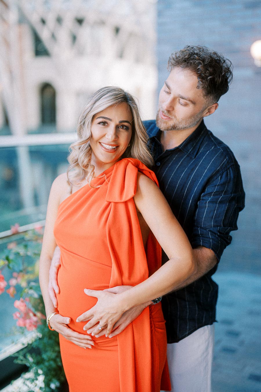 Expectant couple embraces joyfully on a balcony, with the woman wearing an orange dress and the man in a navy shirt, celebrating pregnancy together.