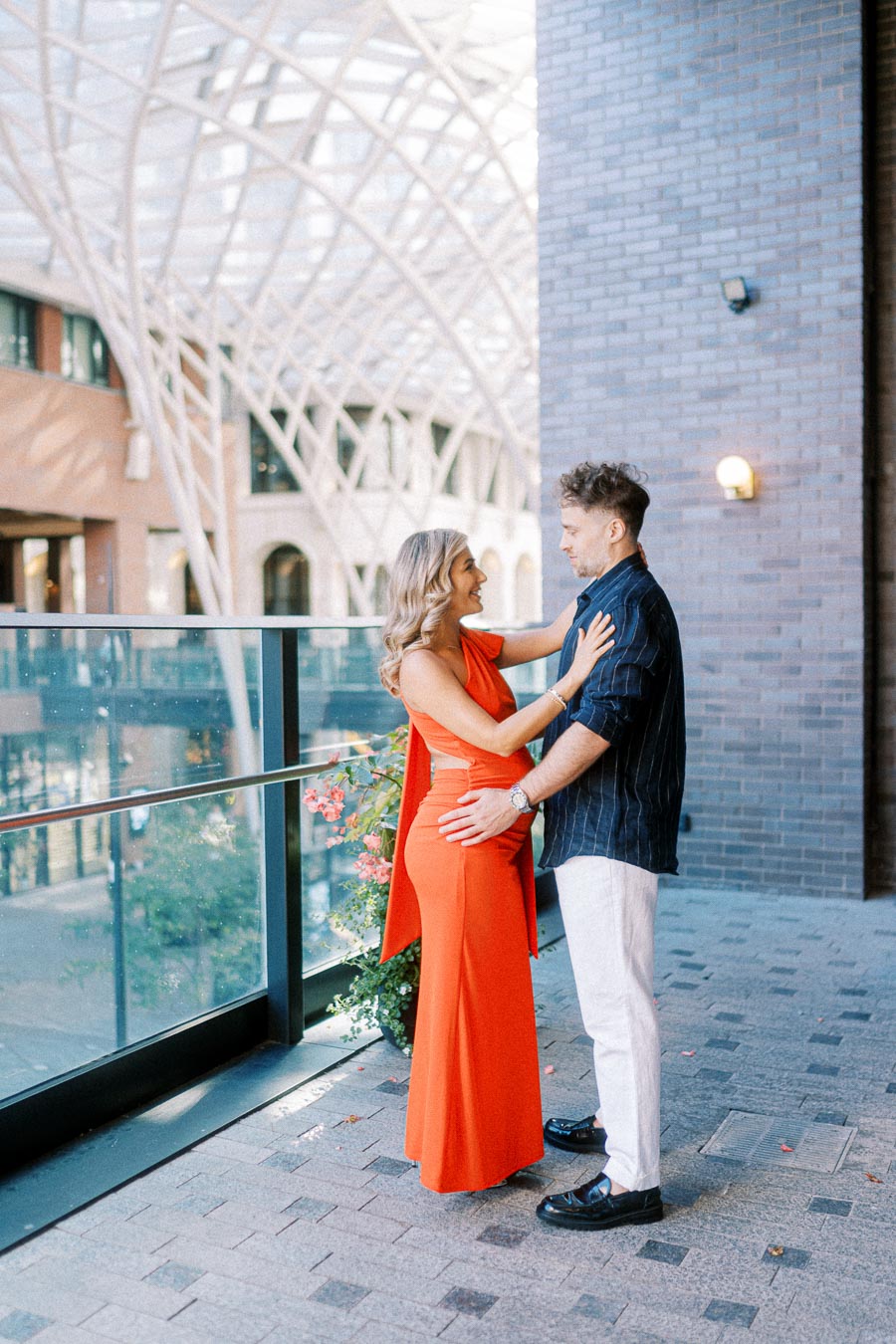A couple embracing on a modern urban balcony with architectural arches in the background, woman in a striking red dress and man in a navy shirt and white pants.