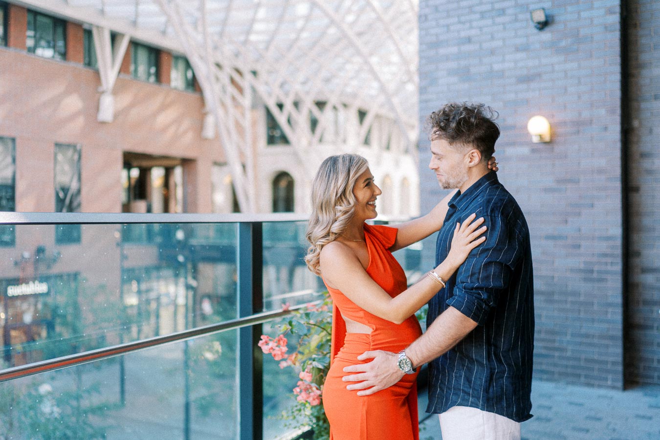 A couple smiling at each other on a modern city balcony, with a glass railing and urban architecture in the background.