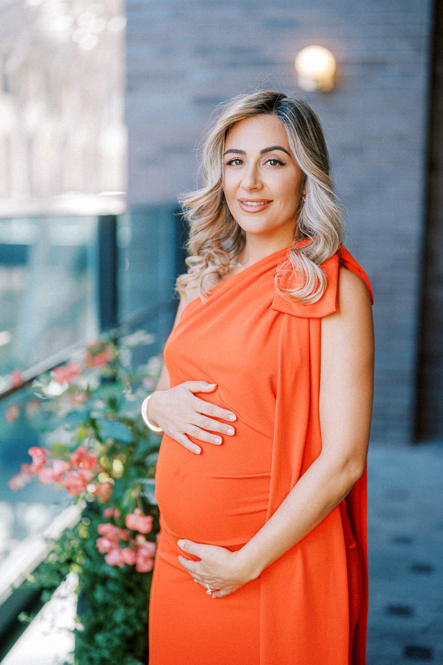 Pregnant woman in a bright orange dress standing outside, gently holding her belly, with pink flowers in the background.