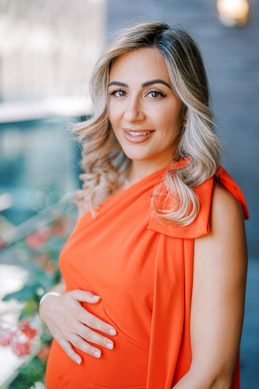 Pregnant woman in a vibrant orange dress smiling and posing outdoors, with a hand gently resting on her belly.