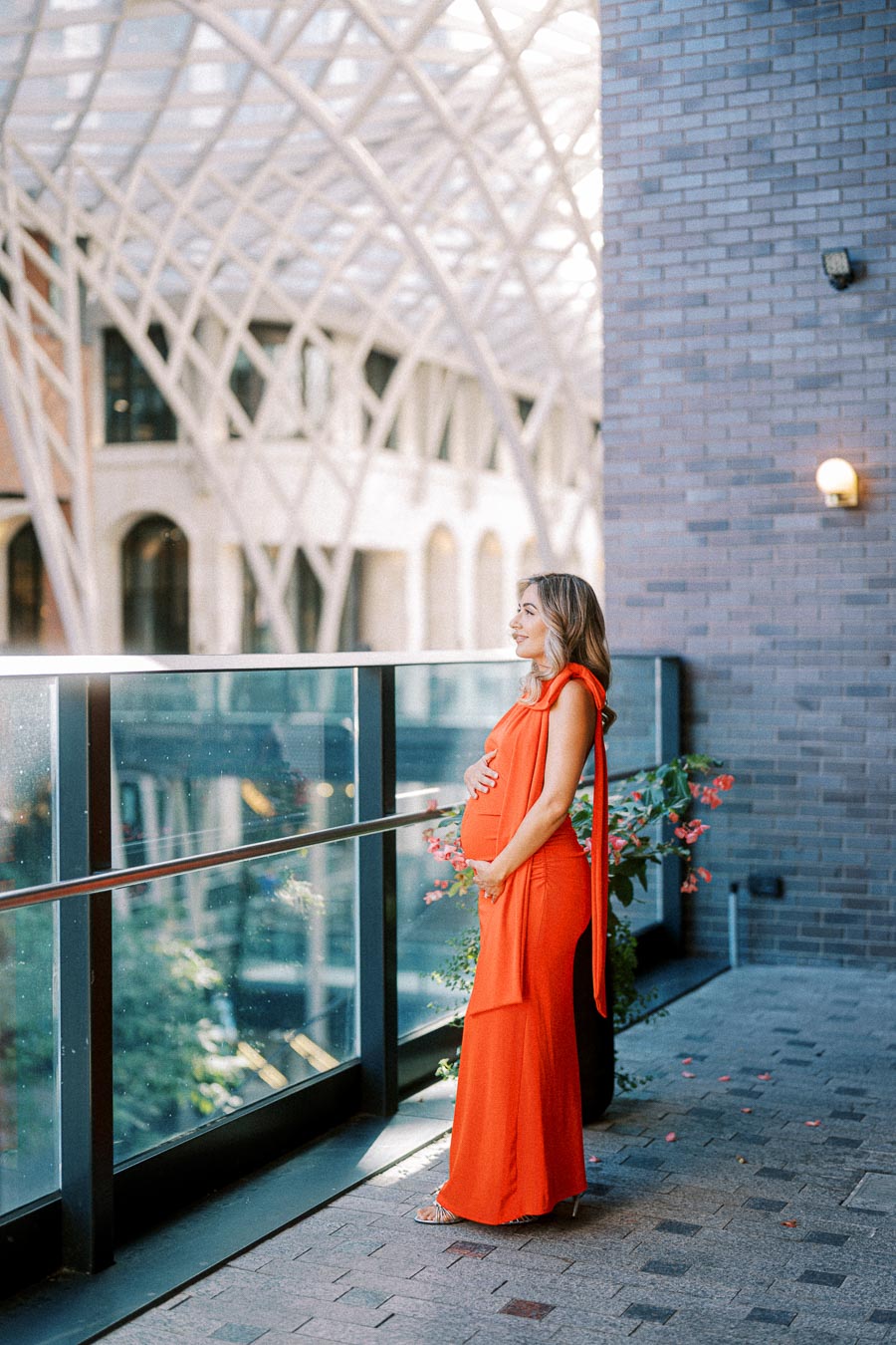 Pregnant woman in an elegant orange dress standing by a glass railing, with an architectural backdrop.