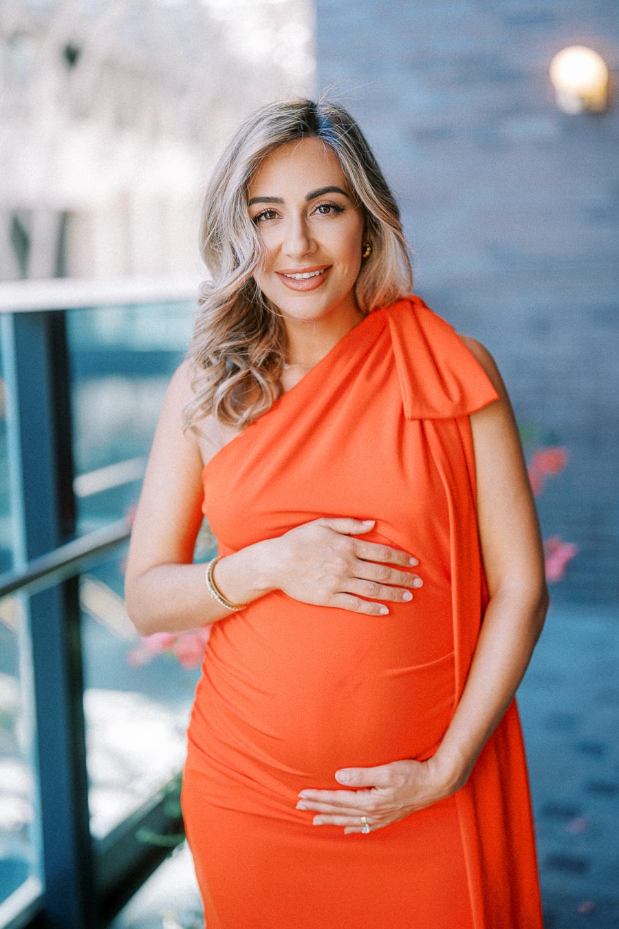 A pregnant woman in an elegant orange dress holds her belly while standing on an outdoor balcony, smiling warmly.