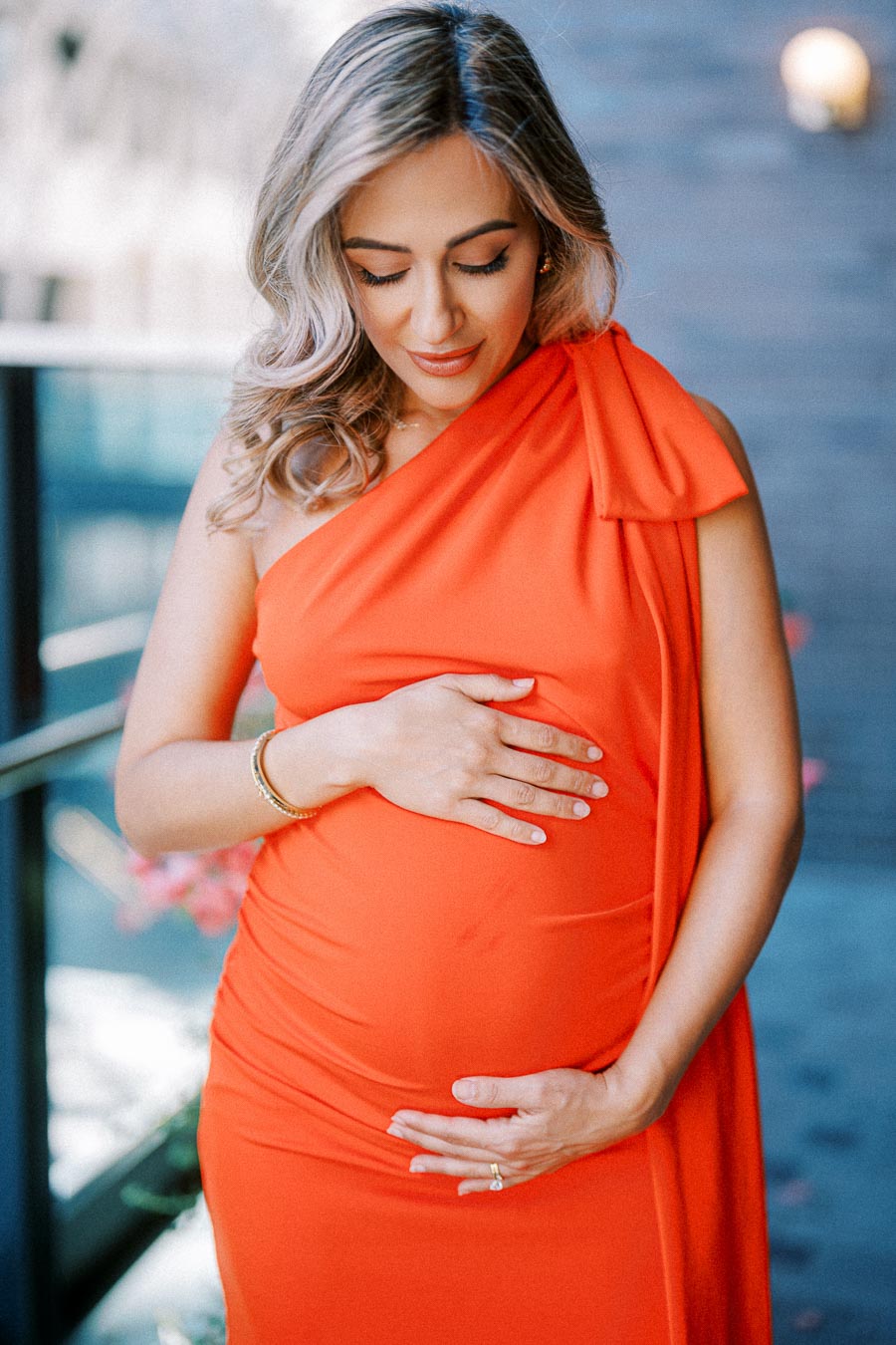 Pregnant woman in an elegant orange dress gently touching her belly while standing outdoors.