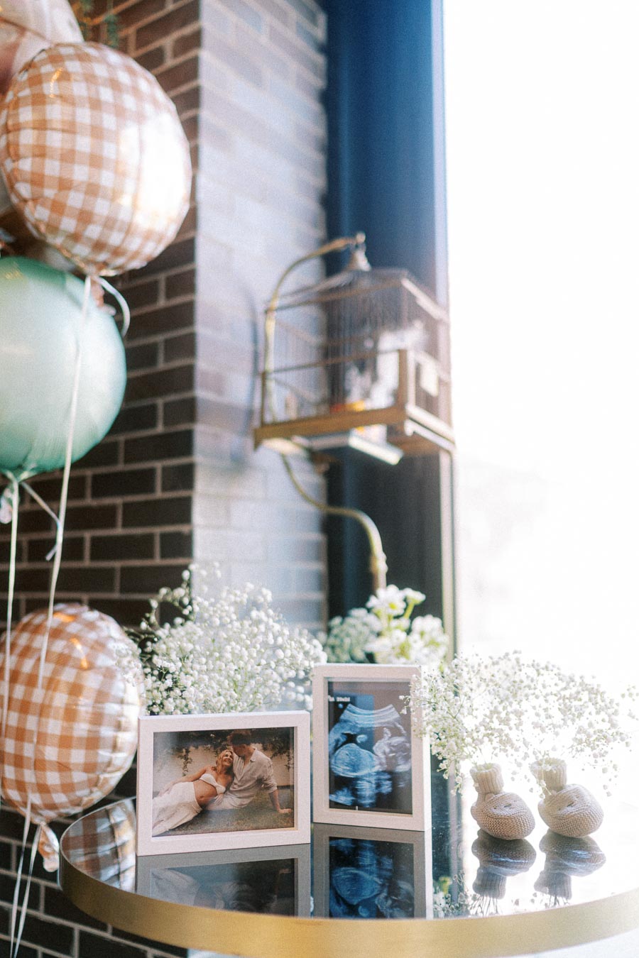 Baby shower decorations on a table featuring plaid balloons, framed ultrasound and couple photographs, baby's breath flowers, and knitted baby booties, with a birdcage and brick wall in the background.