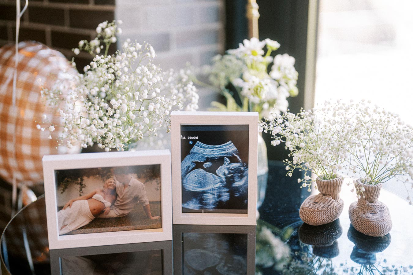 Image featuring a cozy baby shower setup with framed photos, including a maternity shot and an ultrasound image, surrounded by delicate white floral arrangements and tiny knitted baby shoes.