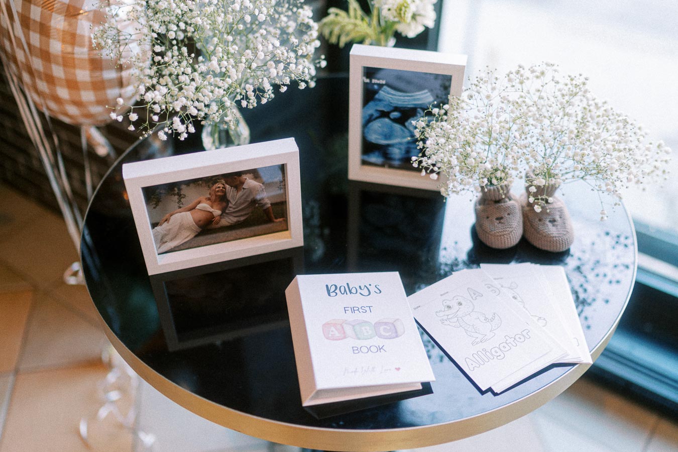 Table setup for a baby shower featuring a baby's ABC book, ultrasound picture, framed maternity photo, baby booties with flowers, and themed coloring cards.
