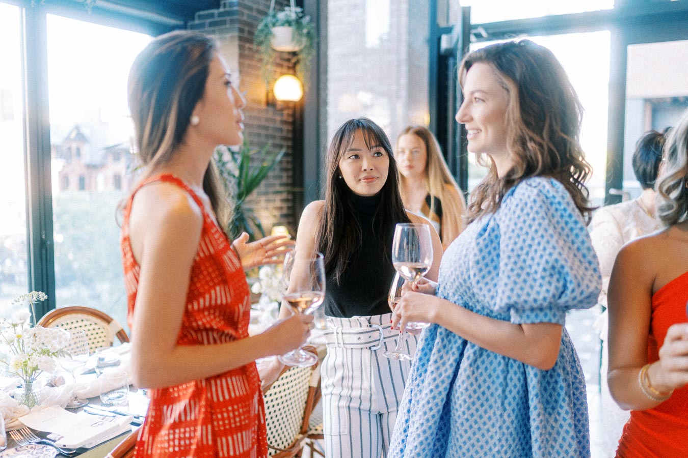 Women socializing at an elegant gathering, holding wine glasses and engaging in conversation, with a stylish, sunlit venue in the background.