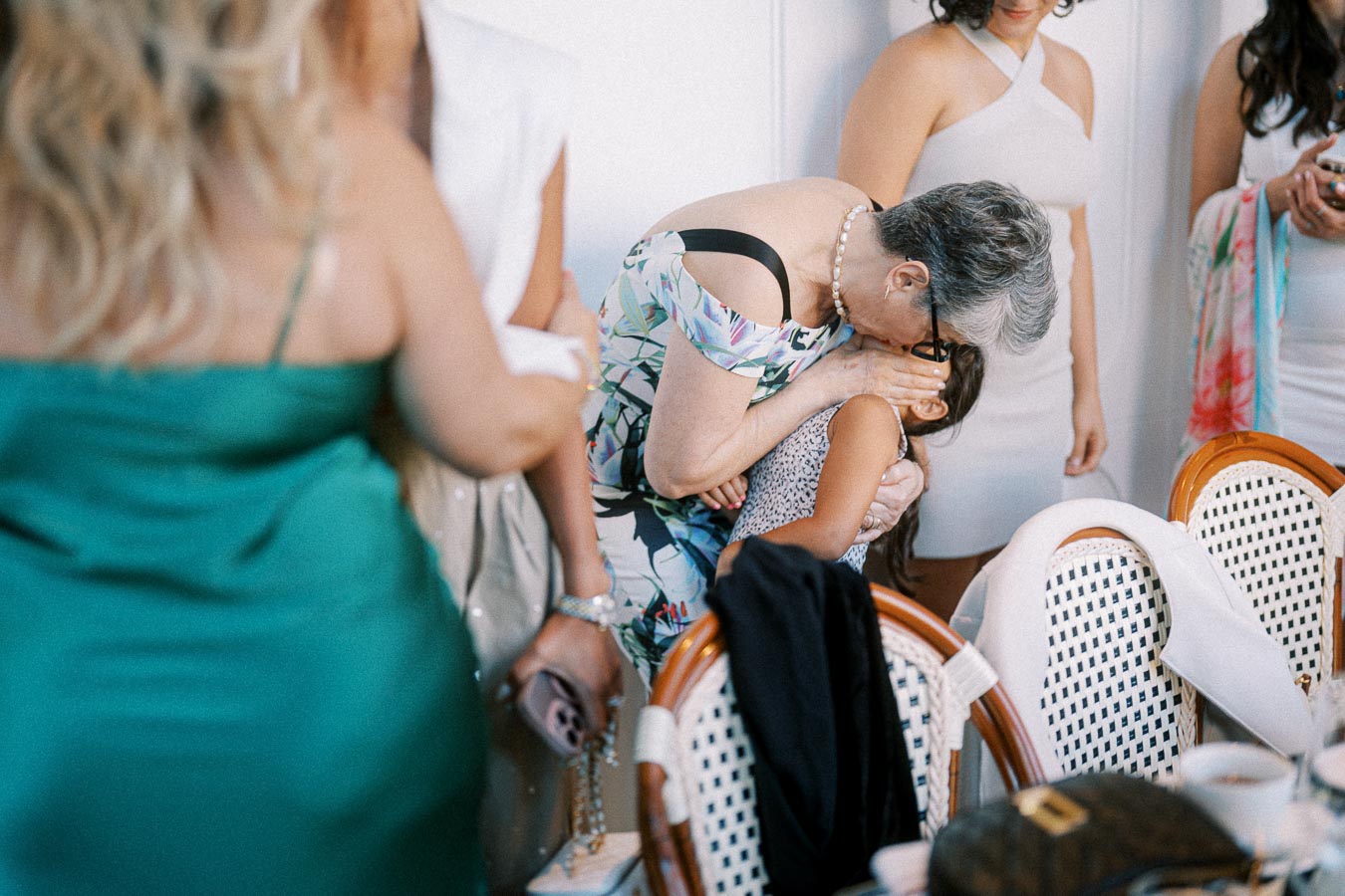 An elderly woman lovingly kisses a young child at a family gathering, surrounded by people in elegant attire, creating a warm and affectionate atmosphere.