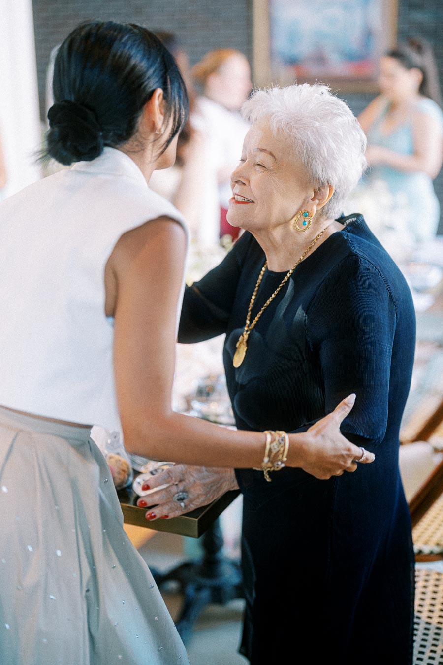 Elderly woman in a navy dress warmly greeting a younger woman in a white blouse and gray skirt during a social gathering.