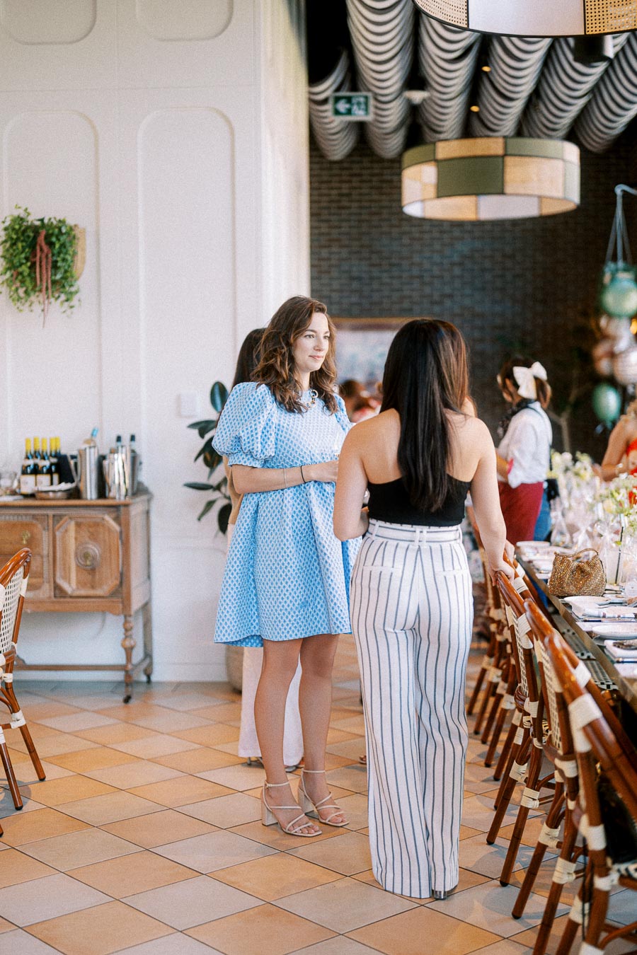Two women chatting at a stylish indoor brunch event, with one wearing a blue dress and the other in a black top and striped pants, surrounded by decor and a set dining table.