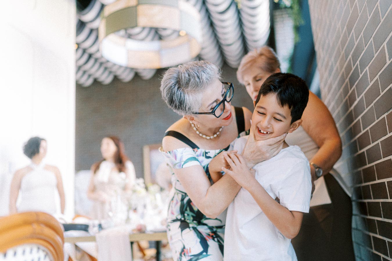 Elderly woman with glasses warmly embraces and smiles at a young boy in a white shirt, surrounded by blurred people in a bright, elegantly decorated room.