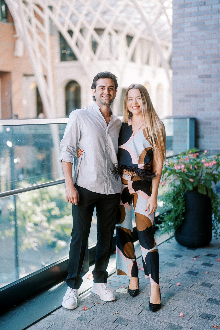 A smiling couple poses together on a modern outdoor patio, with stylish architecture and blooming flowers in the background.