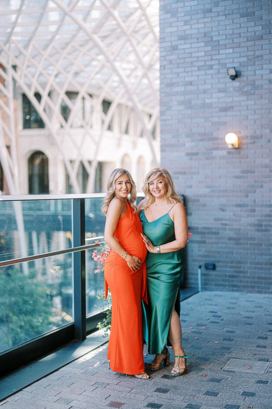 Two women smiling in elegant evening gowns, one in orange and the other in green, standing on a balcony with a modern architectural backdrop.