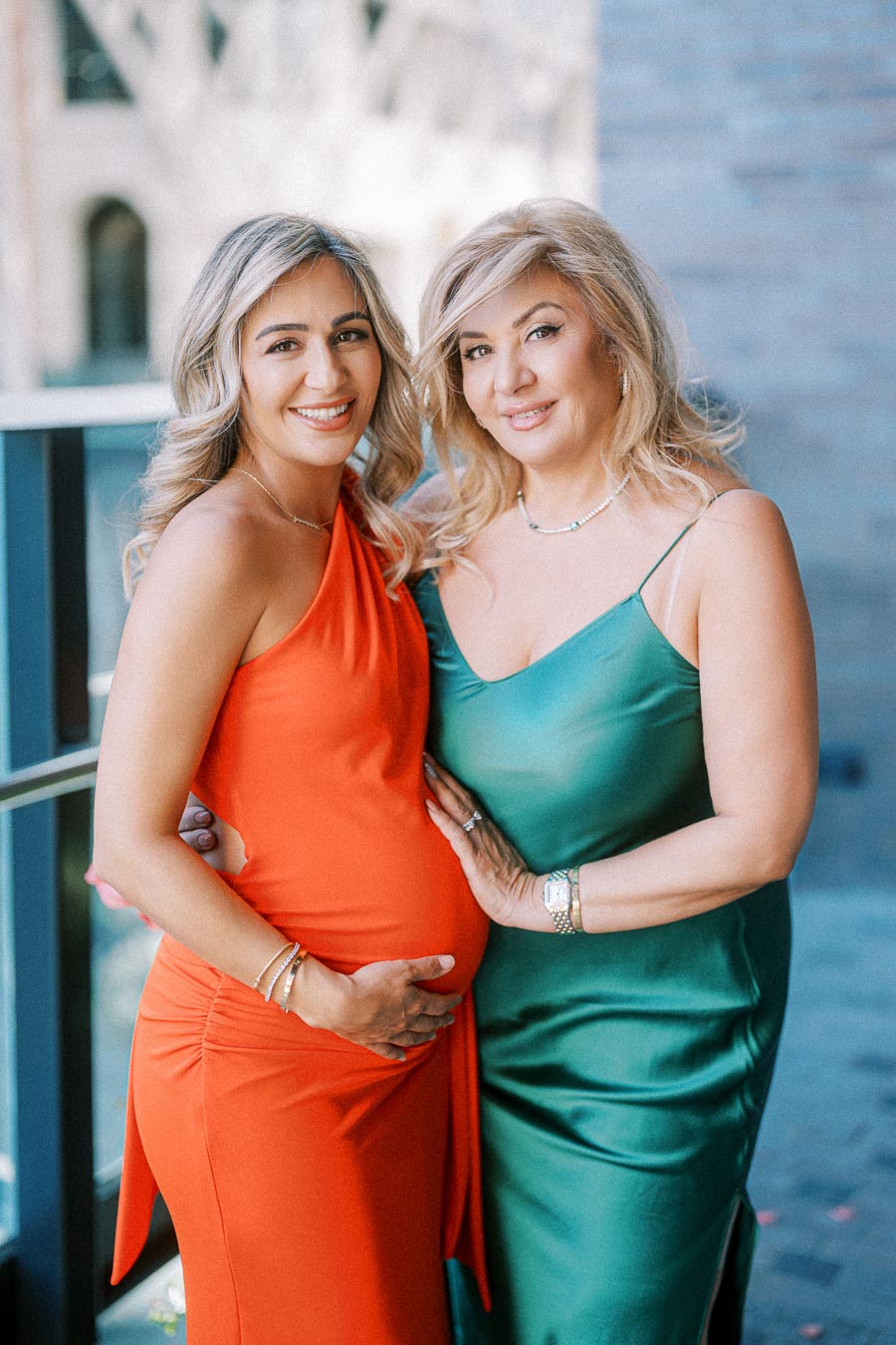 Two women smiling and posing together, with one wearing an orange dress and holding her pregnant belly, and the other in a green dress, standing in a bright, outdoor setting.