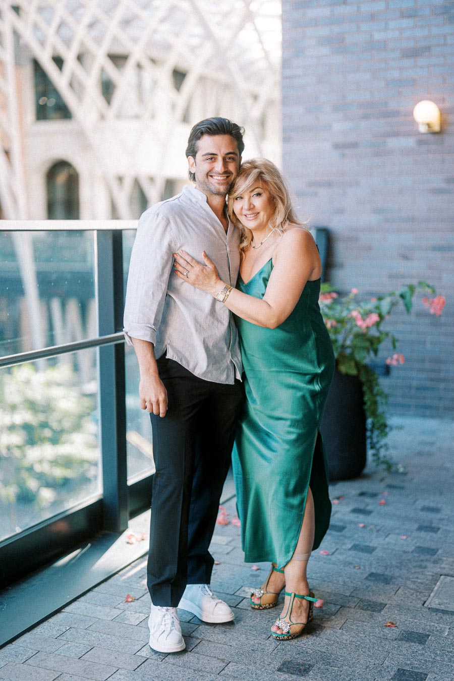 A smiling couple posing together on a modern balcony, with architectural details in the background, adorned in casual elegant attire.