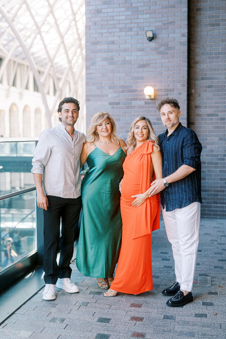 Four people posing together, dressed elegantly; two men flanking two women, with one woman cradling a pregnant belly. The group stands in an architecturally modern, light-filled space, suggesting a celebratory event or family gathering.