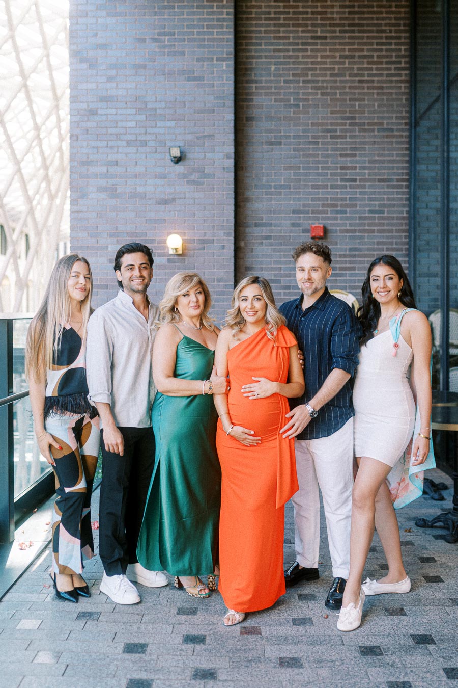 Group portrait of six people smiling together indoors, with one woman in a bright orange dress holding her pregnant belly. The group is styled fashionably, standing against a brick wall with natural light streaming in.