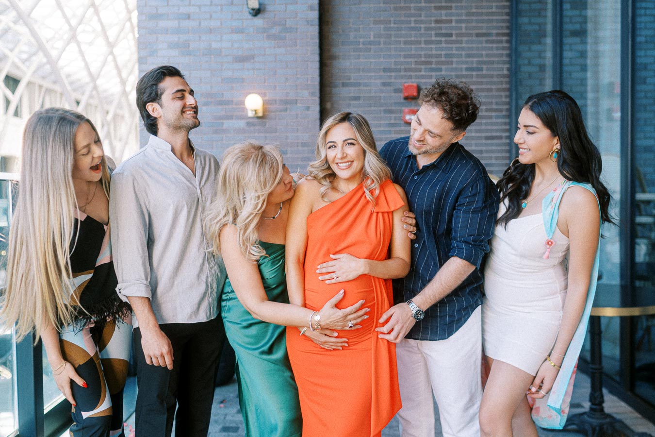 A group of six friends celebrating at a baby shower, with the pregnant woman in the center wearing a bright orange dress, surrounded by smiling guests outdoors against a brick wall background.