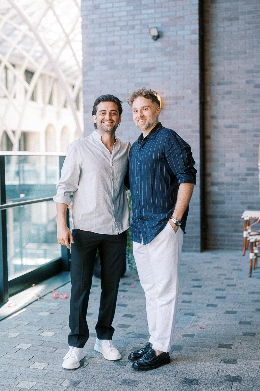 Two men smiling and standing side by side in a modern architectural setting, both dressed in casual, stylish attire.