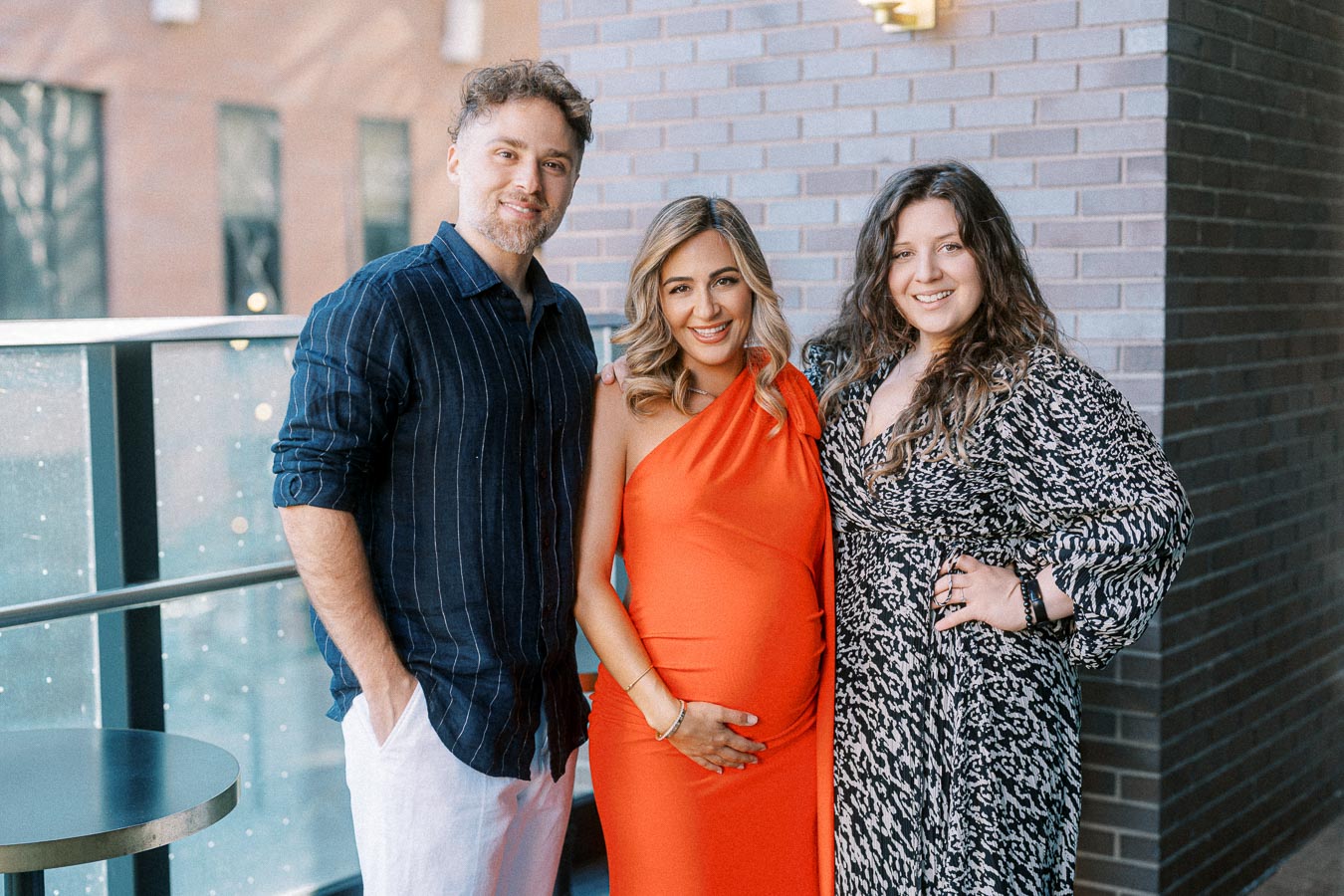 A smiling pregnant woman in an orange dress stands between a man in a striped shirt and a woman in a patterned dress on a balcony, with a brick wall in the background.
