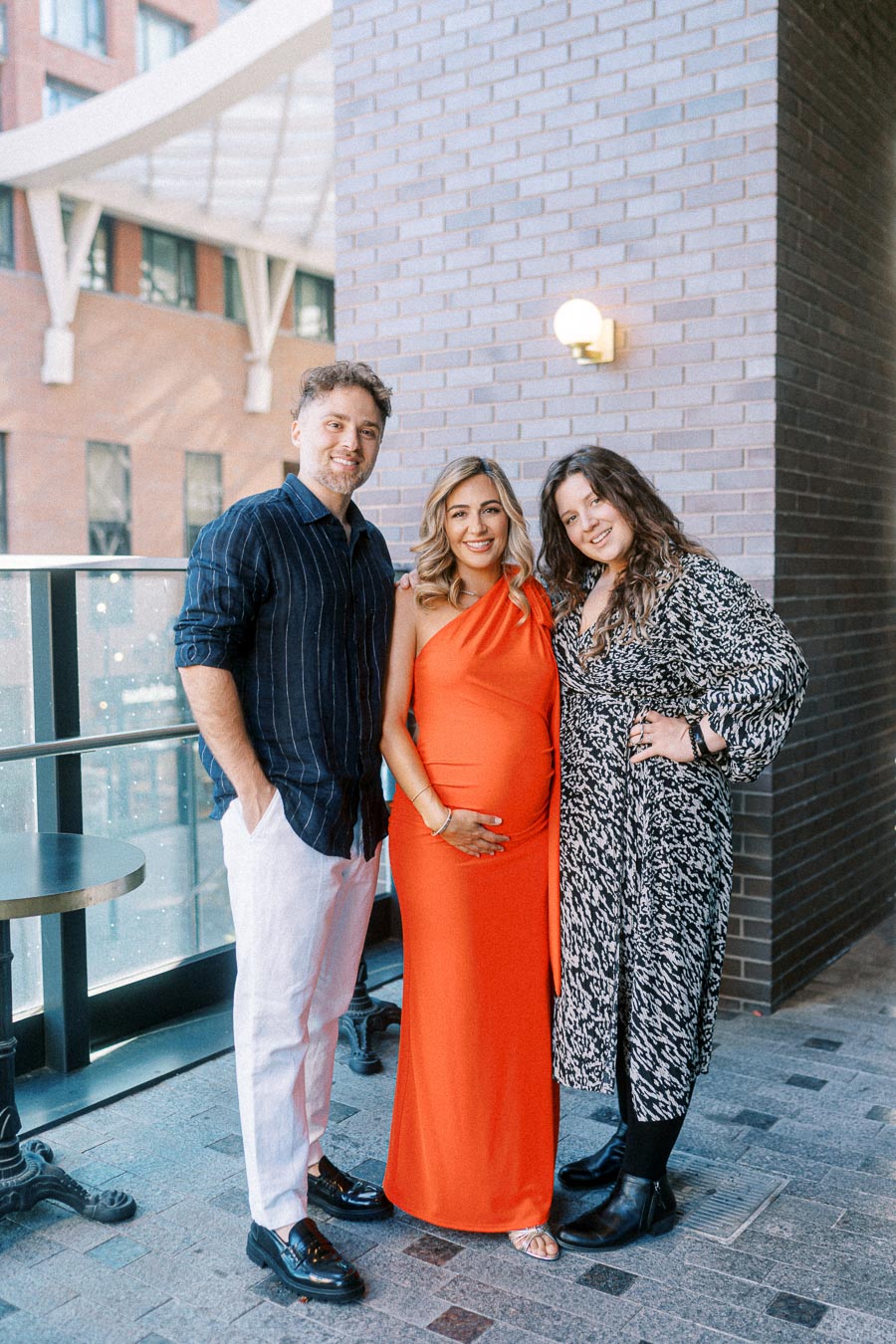 A group photo of three people standing together on a terrace. The person in the middle is wearing an orange dress and appears to be pregnant. They are flanked by a man in a dark shirt and white pants on the left, and a woman in a patterned dress on the right. The backdrop includes a brick wall and a building in the distance.