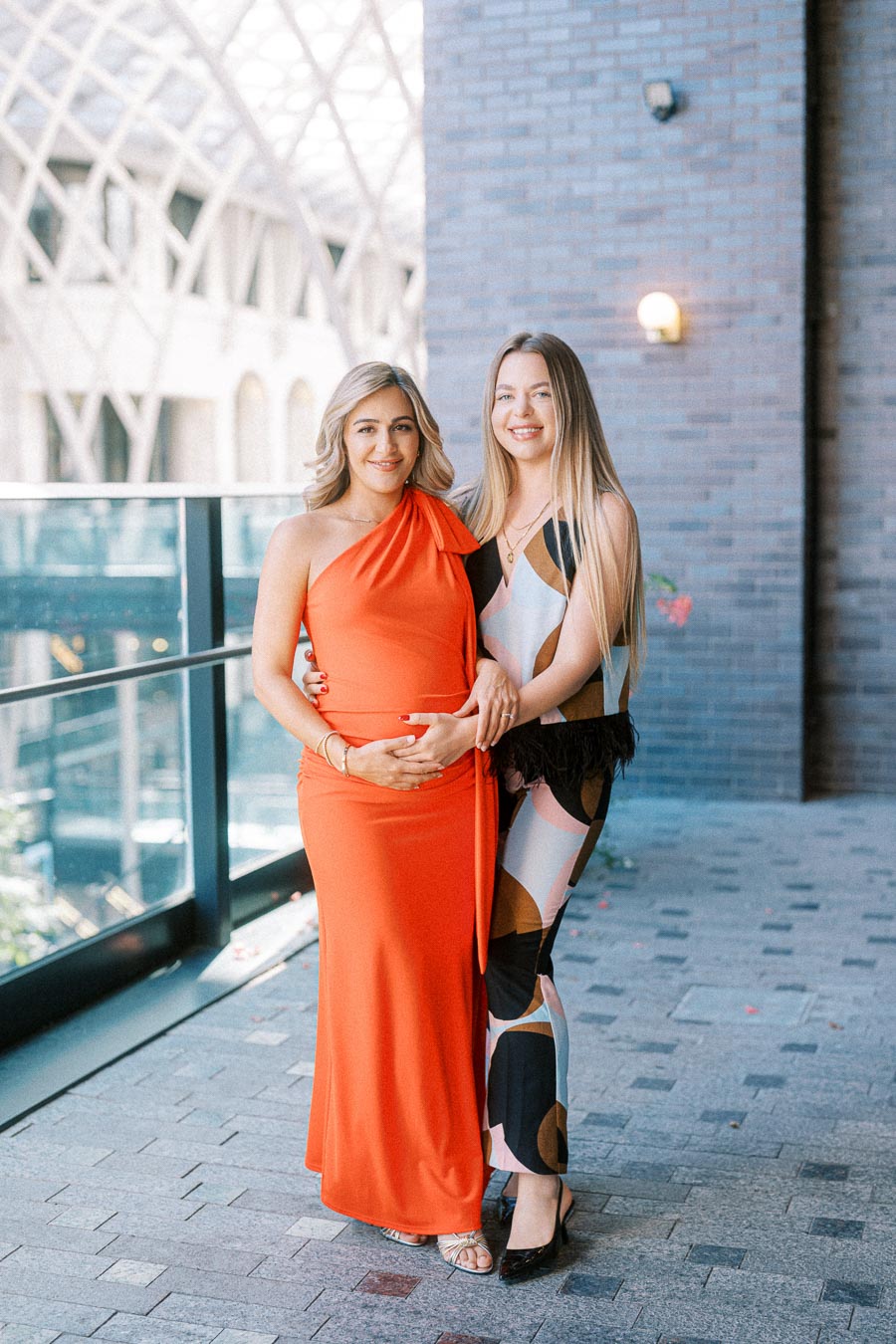 Two women standing together on a balcony. One woman is wearing a bright orange dress, while the other is in a patterned dress. The setting includes a modern architectural background with large glass windows. Both women are smiling and appear to be in a pleasant outdoor environment.