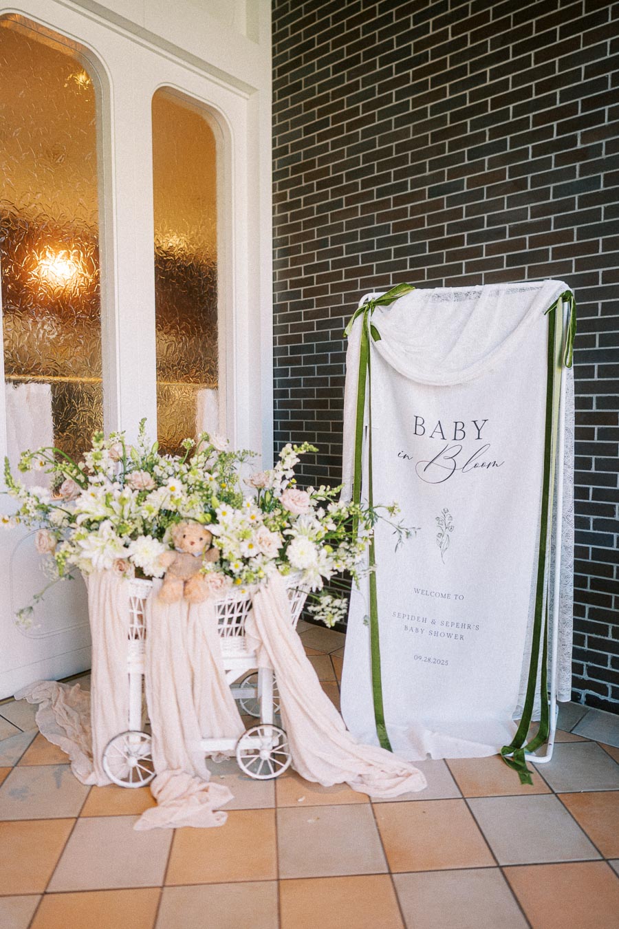 Elegant baby shower decoration featuring a flower-filled pram with a teddy bear, draped in soft fabric, alongside a 'Baby in Bloom' welcome sign, set against a brick wall background.