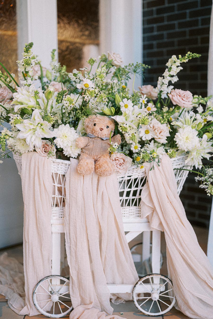 Charming floral arrangement with a teddy bear nestled among white wicker basket filled with pastel flowers and soft fabric drapes, ideal for a vintage or rustic-themed event.