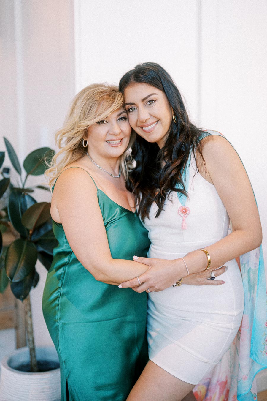 Two women in elegant attire smiling and embracing in a warm, indoor setting, with a decorative plant in the background.